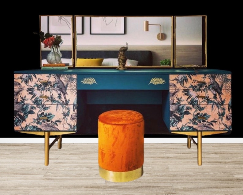 Puede incluir: Una mesa de tocador azul verdoso con tres cajones a cada lado. Los cajones tienen un estampado floral rosa y verde. La mesa tiene detalles dorados y un taburete de terciopelo naranja con ribete dorado. La mesa tiene un espejo de tres paneles con marcos dorados.