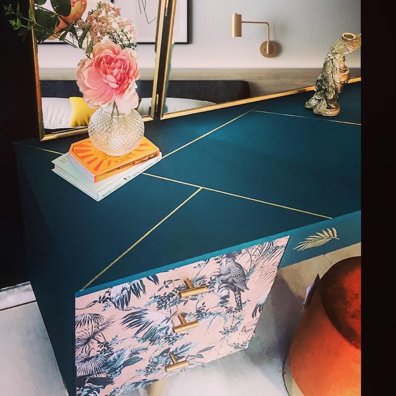 Puede incluir: Una mesa de tocador azul verdoso con detalles dorados y dos cajones. Los cajones est&aacute;n cubiertos con papel tapiz floral rosa y verde con un dise&ntilde;o de p&aacute;jaro. La mesa tiene un borde dorado alrededor de los bordes y un dise&ntilde;o de hoja de oro en la parte superior.