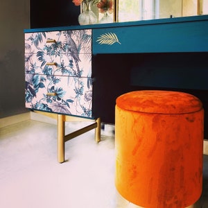Puede incluir: Un tocador azul teal con un caj&oacute;n con estampado floral y patas doradas. Un otomano de terciopelo naranja brillante se encuentra frente al tocador.