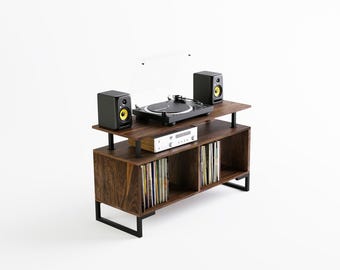 May include: A dark brown wooden record player stand with black metal legs. The stand has two shelves for storing records and a top shelf for a turntable, amplifier, and speakers. The turntable is black with a clear dust cover. The speakers are black with yellow accents.
