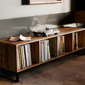 Può includere: Un mobile in legno per giradischi con gambe in metallo nero. Il mobile ha cinque ripiani per riporre dischi in vinile. Un giradischi è sul ripiano superiore e una rivista è sul secondo ripiano.