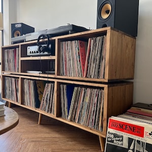 Op de afbeelding: Een houten draaitafelstandaard met twee planken, elk met rijen vinylplaten. De standaard heeft een draaitafel bovenop en twee luidsprekers aan weerszijden. De standaard is afgewerkt met een natuurlijke houtkleur en heeft een rustieke uitstraling. De standaard staat op een houten vloer.