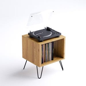 Może przedstawiać: Drewniany stojak na gramofon z czarnymi metalowymi nogami. Stojak ma półkę do przechowywania płyt i górną powierzchnię na gramofon. Gramofon ma przezroczystą osłonę przeciwkurzową.
