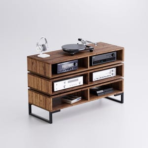 Könnte beinhalten: Ein Unterhaltungszentrum aus Holz mit schwarzen Metallbeinen. Das Zentrum hat fünf Regale, auf denen jeweils eine andere Audiokomponente steht, darunter ein Plattenspieler, ein Verstärker und ein Receiver. Ein Paar weiße Kopfhörer sitzt oben auf dem Zentrum.