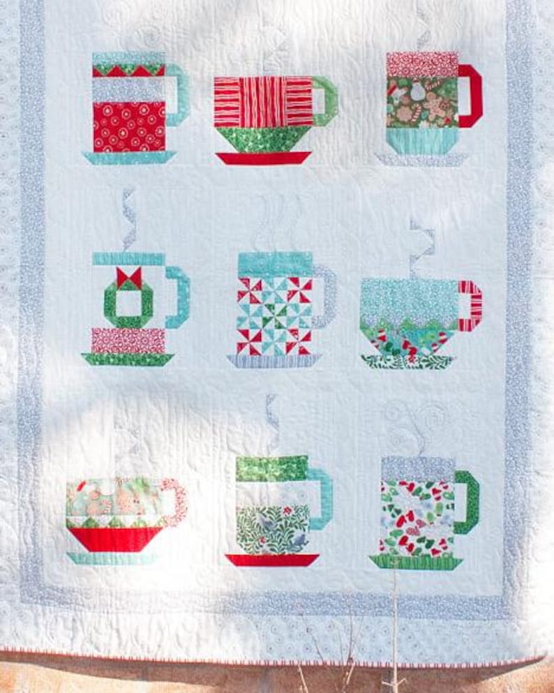 May include: A white quilt with a patchwork design of nine colorful mugs. Each mug is made of different fabrics and patterns, including red, green, blue, and white. The mugs are arranged in a 3x3 grid.