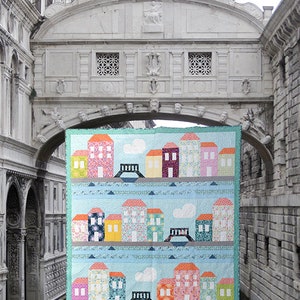 May include: A colourful quilt depicting a cityscape with houses, bridges, and clouds. The quilt is displayed against a backdrop of a stone archway.