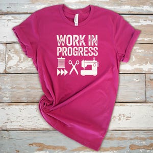 May include: A magenta t-shirt with the text "WORK IN PROGRESS" in white, along with white illustrations of a spool of thread, scissors, and a sewing machine. The shirt is folded, showing the rolled-up sleeves.
