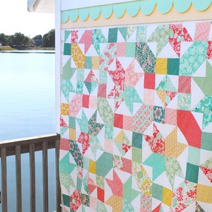 May include: A patchwork quilt with star and square patterns in red, green, yellow, and teal. The quilt is displayed outdoors, hanging on a white wall. The quilt's design is set against a white background. The quilt is displayed near a body of water.