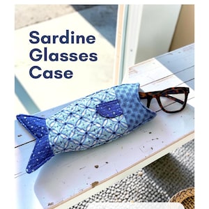 Può includere: Un astuccio per occhiali in tessuto trapuntato a forma di pesce con motivi blu e azzurri. L'astuccio presenta una piccola pinna e una coda, con un paio di occhiali dalla montatura scura all'interno. Il testo recita Kate Spain, Sardine Glasses Case, KS 2603, PDF Sewing Pattern.
