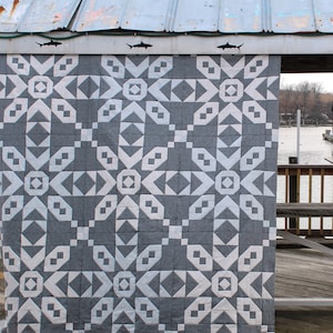 Peut inclure: Une courtepointe à motifs géométriques gris et blancs est suspendue à une structure métallique patinée. Le motif de la courtepointe présente des formes répétitives de losanges, de carrés et de chevrons. Le fond est gris uni et les formes sont blanches. La structure a des silhouettes de requins.