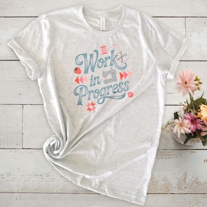 Op de afbeelding: Lichtgrijze t-shirt met opgerolde mouwen met de tekst "Work in Progress" in blauw schrift. Het ontwerp bevat illustraties met naaithema's, zoals een naaimachine, een schaar en een klos garen. Een klein boeket bloemen staat rechts.