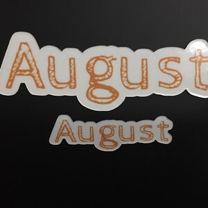 May include: Two white stickers with the word "August" written in orange. The top sticker is larger and has a more stylized font.