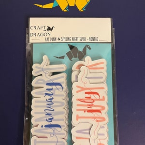 Puede incluir: Un paquete de pegatinas con los meses del a&ntilde;o en fuente cursiva. Las pegatinas son de colores pastel, con la palabra "Enero" en azul y "Julio" en rojo. El paquete est&aacute; etiquetado como "Craft Dragon" y "Rae Dunn & Spelling Night Swirl - Months". Un drag&oacute;n de origami amarillo en la parte superior.