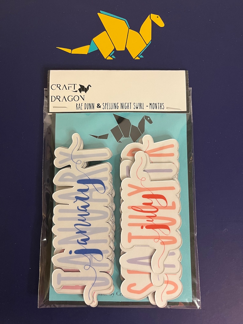 Puede incluir: Un paquete de pegatinas con los meses del a&ntilde;o en fuente cursiva. Las pegatinas son de colores pastel, con la palabra "Enero" en azul y "Julio" en rojo. El paquete est&aacute; etiquetado como "Craft Dragon" y "Rae Dunn & Spelling Night Swirl - Months". Un drag&oacute;n de origami amarillo en la parte superior.