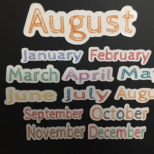 May include: A set of twelve colorful month stickers with a handwritten font. The stickers are on a black background. The months are January, February, March, April, May, June, July, August, September, October, November, and December.