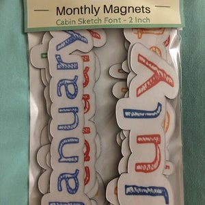 May include: A set of 2-inch magnetic letters in a cabin sketch font. The letters are in various colors, including blue, red, orange, and green. The letters are arranged in a pile, with the letters "January" and "July" visible.
