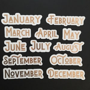 May include: Twelve white stickers with gold lettering, each featuring a different month of the year. The stickers are arranged in three rows of four stickers each.