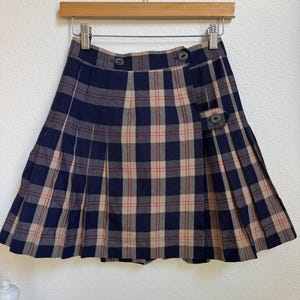 May include: A pleated plaid skirt with a navy blue, beige, and red plaid pattern. The skirt has a button closure and a flap pocket on the side.
