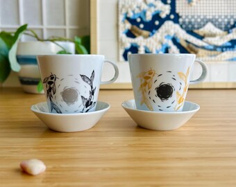 May include: Two white ceramic teacups with saucers. One cup has a black and white design of two fish swimming in a circle. The other cup has a gold and white design of two fish swimming in a circle.