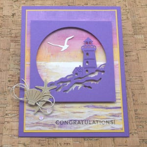 May include: A rectangular greeting card with a purple border and a sunset-colored background. A purple die-cut overlay features a lighthouse and a seagull. A tan seashell and starfish are attached with twine. The word CONGRATULATIONS is printed below.