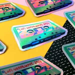 May include: A holographic sticker of a cassette tape with the text "LOFI-VAPOR WAVE-24/7" on it. The sticker is green, pink, and blue.