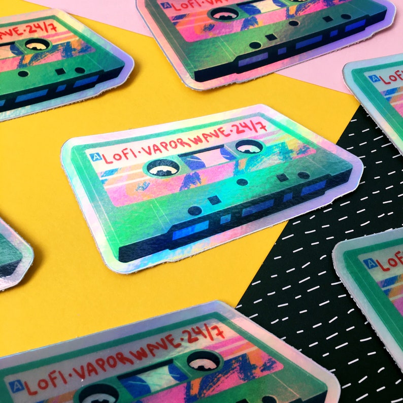 May include: A holographic sticker of a cassette tape with the text "LOFI-VAPOR WAVE-24/7" on it. The sticker is green, pink, and blue.
