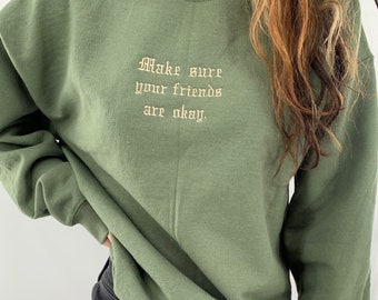 Puede incluir: Una sudadera verde con el texto "Make sure your friends are okay." bordado en la parte delantera. La sudadera se combina con unos pantalones cortos de mezclilla negros.