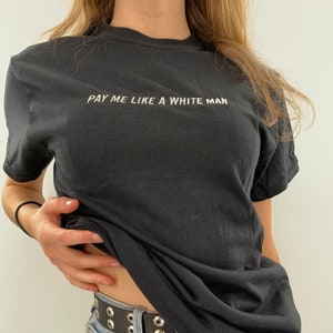 Puede incluir: Una camiseta negra con el texto "PAY ME LIKE A WHITE MAN" en letras blancas. La camiseta la lleva puesta una persona con un cinturón negro tachonado y unos vaqueros azules.