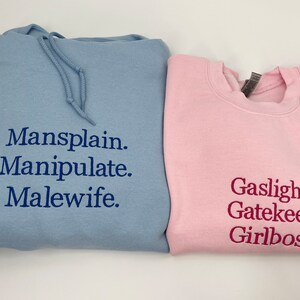 Puede incluir: Dos sudaderas con texto impreso. La sudadera azul tiene el texto "Mansplain. Manipulate. Malewife." impreso en ella. La sudadera rosa tiene el texto "Gaslight. Gatekeep. Girlboss." impreso en ella.