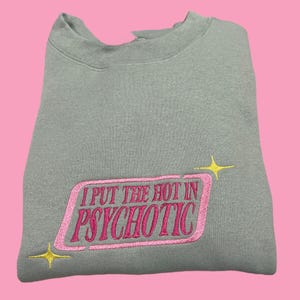 A gray sweatshirt with a pink and yellow embroidered phrase that reads "I PUT THE HOT IN PSYCHOTIC" on it.