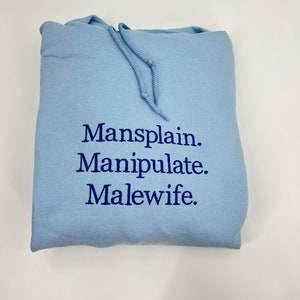 Puede incluir: Sudadera con capucha azul claro con el texto "Mansplain. Manipulate. Malewife." bordado en azul oscuro.