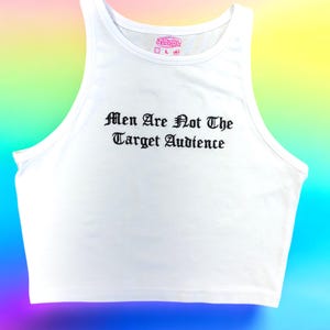 Puede incluir: Camiseta sin mangas blanca con el texto "Men Are Not The Target Audience" en fuente gótica negra. La camiseta tiene un diseño de espalda nadadora y una pequeña etiqueta rosa. El fondo es un degradado arcoíris.