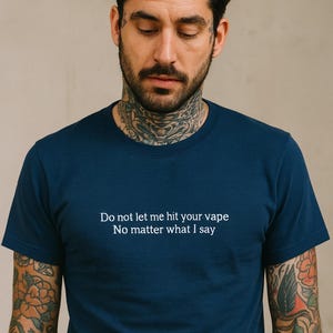 A man with tattoos on his arms wearing a blue t-shirt with a humorous message printed on it.