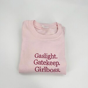 Puede incluir: Sudadera rosa con el texto "Gaslight. Gatekeep. Girlboss." impreso en granate.