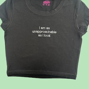 May include: Black crop top with the embroidered text "I am as unapproachable as I look." The shirt has a crew neck and short sleeves. A small pink tag is visible near the neckline.