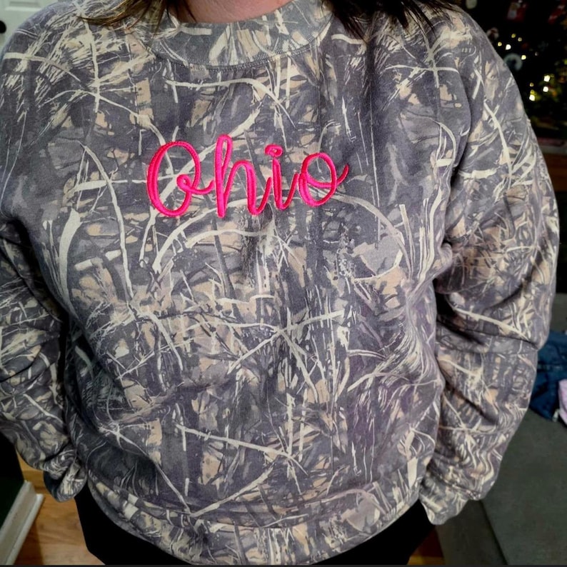 A person wearing a camouflage-patterned sweatshirt with the word "Ohio" embroidered on it in pink.