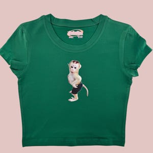 May include: Green short-sleeved t-shirt with a small monkey graphic wearing black shorts. The shirt has a pink label that says "SMALL" and "MADE IN CHINA".