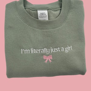 May include: A sage green sweatshirt with the text "I'm literally just a girl" embroidered in white with a pink bow underneath.