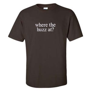 Op de afbeelding: Een donkerbruin T-shirt met de tekst "where the huzz at?" in wit gedrukt op de voorkant.