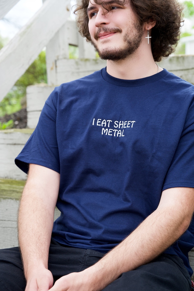 Op de afbeelding: Een marineblauw t-shirt met de tekst "I EAT SHEET METAL" in wit gedrukt op de voorkant.