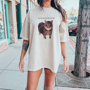 Puede incluir: Una mujer lleva una camiseta beige de gran tamaño con un gato atigrado marrón estampado en la parte delantera. La camiseta tiene el texto "oo ee aa oo ee ee aa" impreso sobre el gato.