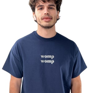 Può includere: Una t-shirt blu navy con la parola "womp" stampata due volte in bianco sul davanti.