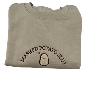 Puede incluir: Una sudadera beige con el texto "MASHED POTATO SLUT" bordado en marrón. Una pequeña patata de dibujos animados con un corazón rojo encima está bordada en el centro del texto.