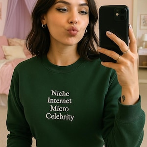 A young woman in a green sweater and denim shorts is taking a selfie with a black smartphone, while a pink canopy can be seen in the background.
