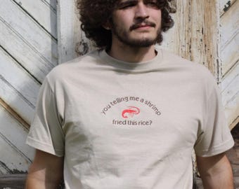 May include: A person wearing a light beige t-shirt with a red shrimp graphic and the text "you telling me a shrimp fried this rice?"