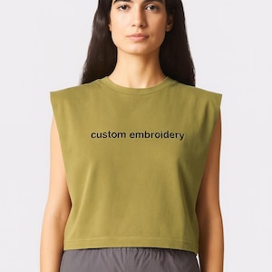 May include: Olive green sleeveless crop top with the text "custom embroidery" in black. The model is wearing gray pants. The top has a crew neck and a relaxed fit. The background is plain white.
