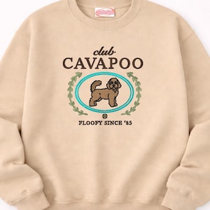 May include: Beige crew neck sweatshirt featuring the embroidered text "club CAVAPOO" above an oval design with a brown dog illustration. The design is surrounded by a teal border and a green wreath. The text "FLOOFY SINCE '85" is below the oval.