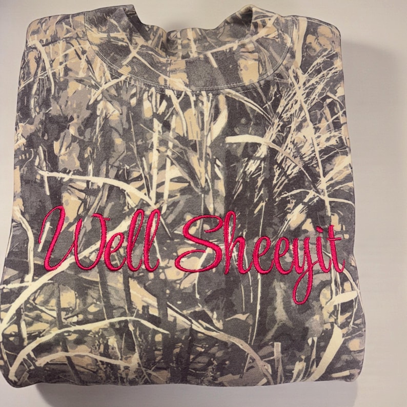 A camouflage-patterned t-shirt with the text "Well Sheeyit" embroidered on it.