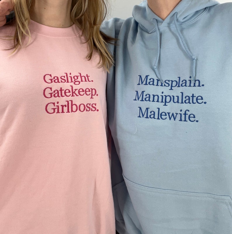 Puede incluir: Dos sudaderas con texto impreso en la parte delantera. La sudadera rosa dice "Gaslight. Gatekeep. Girlboss." La sudadera azul dice "Mansplain. Manipulate. Malewife."