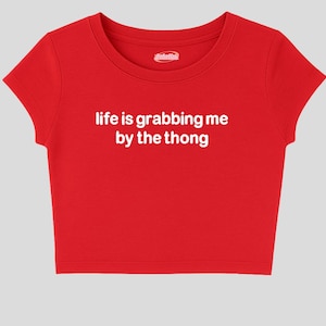 Puede incluir: Una camiseta corta roja brillante con cuello redondo y mangas cortas. La camiseta tiene el texto blanco "life is grabbing me by the thong" en la parte delantera. Un pequeño logotipo blanco es visible cerca del cuello.